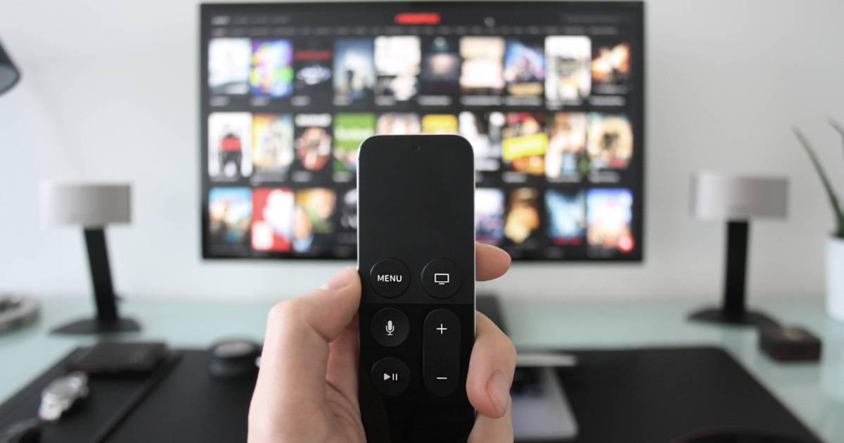 A hand holds a TV remote, pointing at a large screen displaying a streaming service interface with various thumbnails. This depiction highlights the potential for maximizing OTT video monetization.